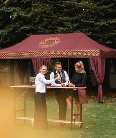 Elegant folding gazebo 8 x 4 m burgundy with gold decorations and decorative pole-covering curtains. The gazebo stands in a meadow and is used for the reception of an elegant event while people drink sparkling wine served by a waitress